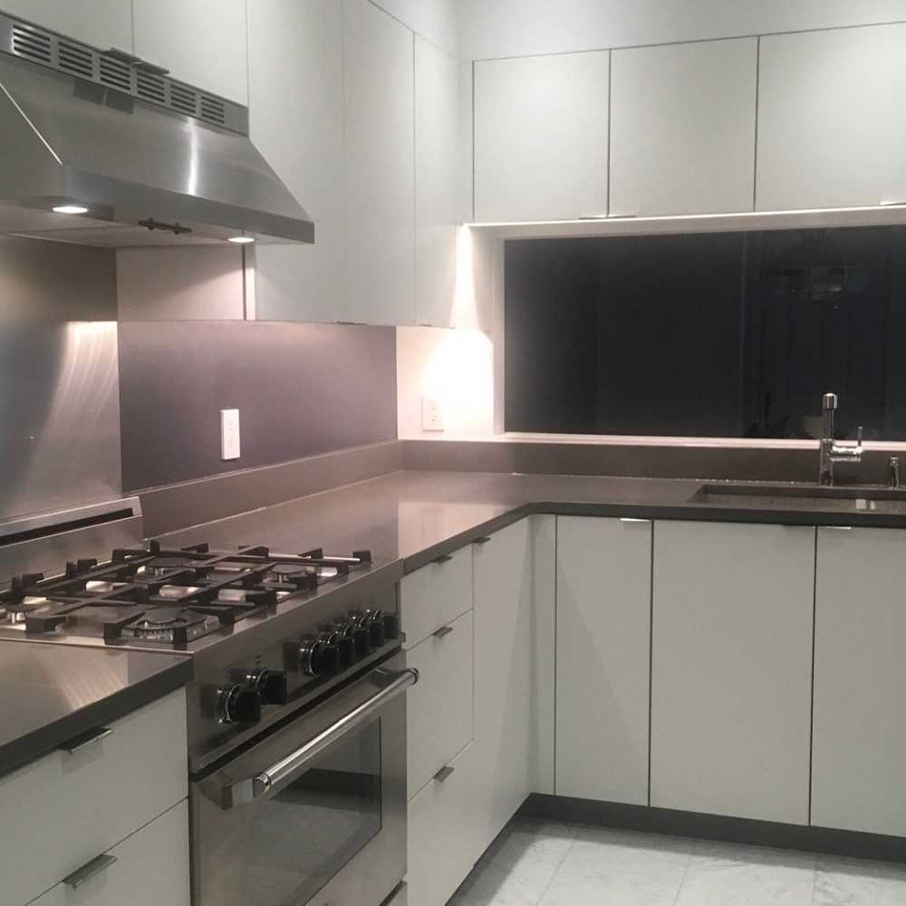 White flat-panel cabinets, a stainless hood and backsplash, a commercial-style range and a full-width pass-through lit by LED.