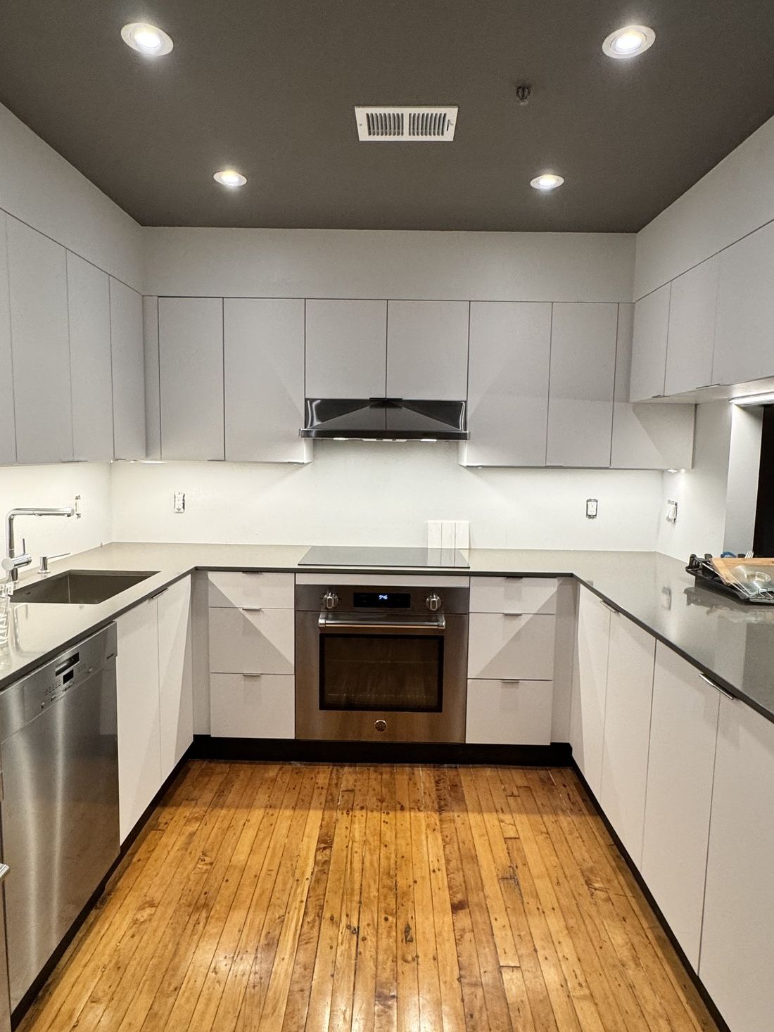 A U-shaped kitchen with flat-panel white cabinets, dark counters, a cooktop with wall oven below, and the original wood floor.