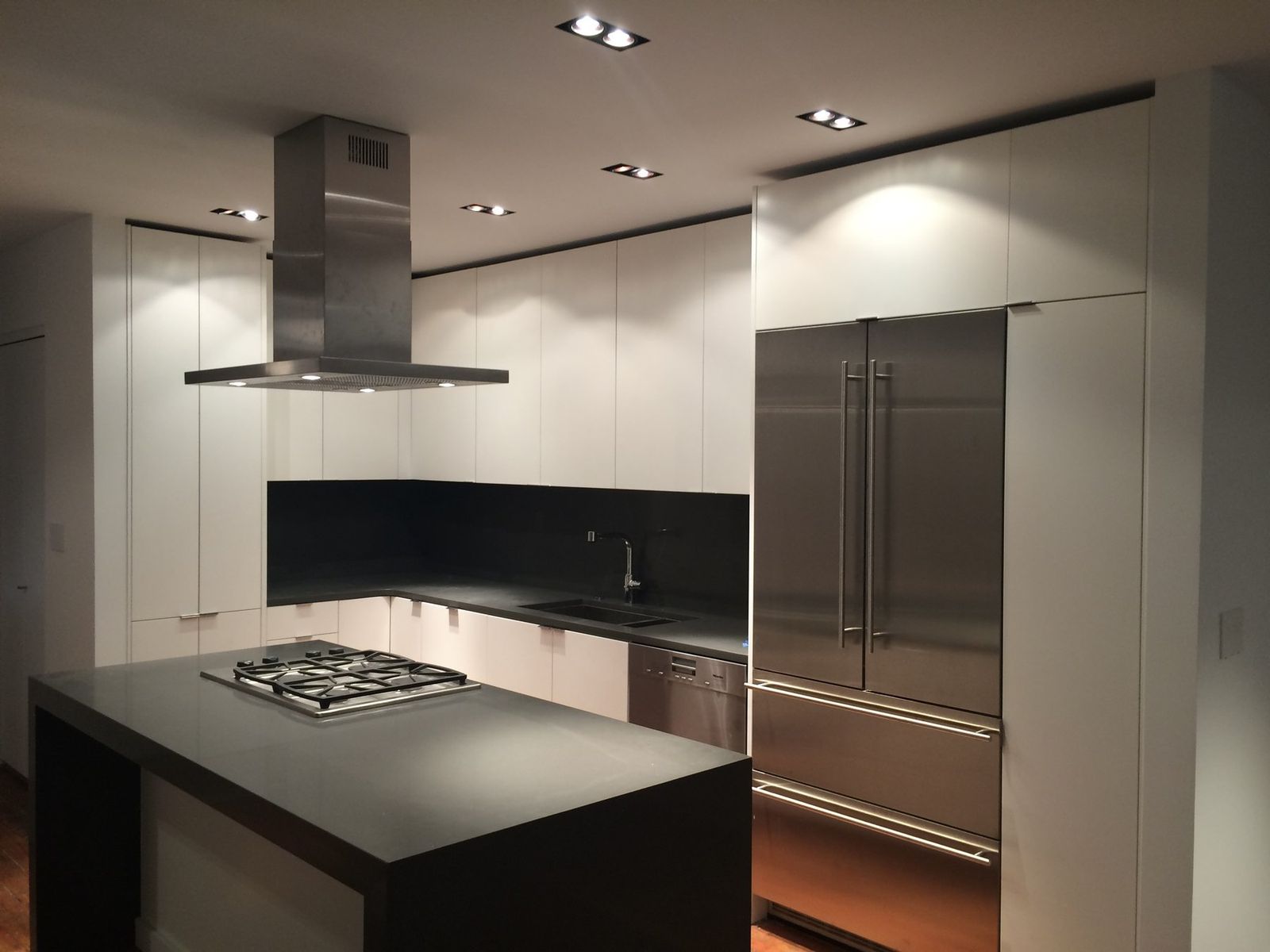 A white and charcoal kitchen with a waterfall island, a cooktop, an island hood and a tall built-in refrigerator wall.